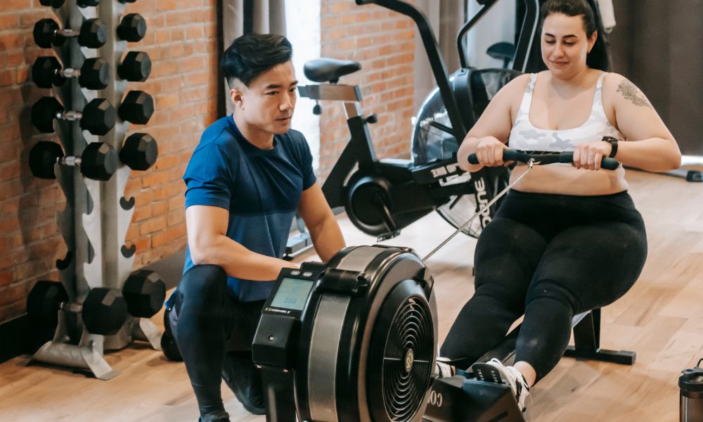 Personal trainer orientando a cliente obesa em um ergômetro simulador de remo.