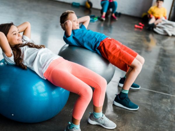 crianças realizando abdominal na bola suíça em uma academia com área kids