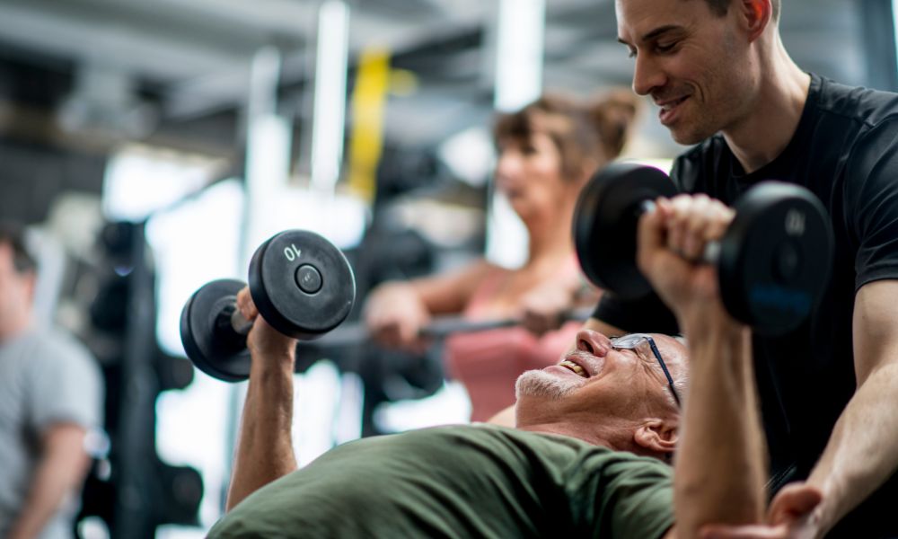 Personal trainer dando assistência a um aluno dentro de uma sala de musculação.